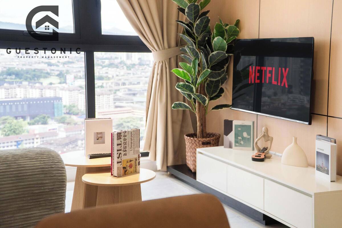 Una habitación de hotel moderna con un ventanal grande, un televisor de pantalla plana mostrando 'NETFLIX', una planta en maceta, una mesita con libros y un mueble blanco.