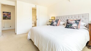 A bedroom with a large bed, a nightstand, a lamp, a painting, and an open door leading to a bathroom. - Old Railway Close (Lechlade)