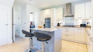 A modern kitchen with white cabinetry, a built-in oven, a stainless steel range hood, and a bar area with two stools. - Old Railway Close (Lechlade)