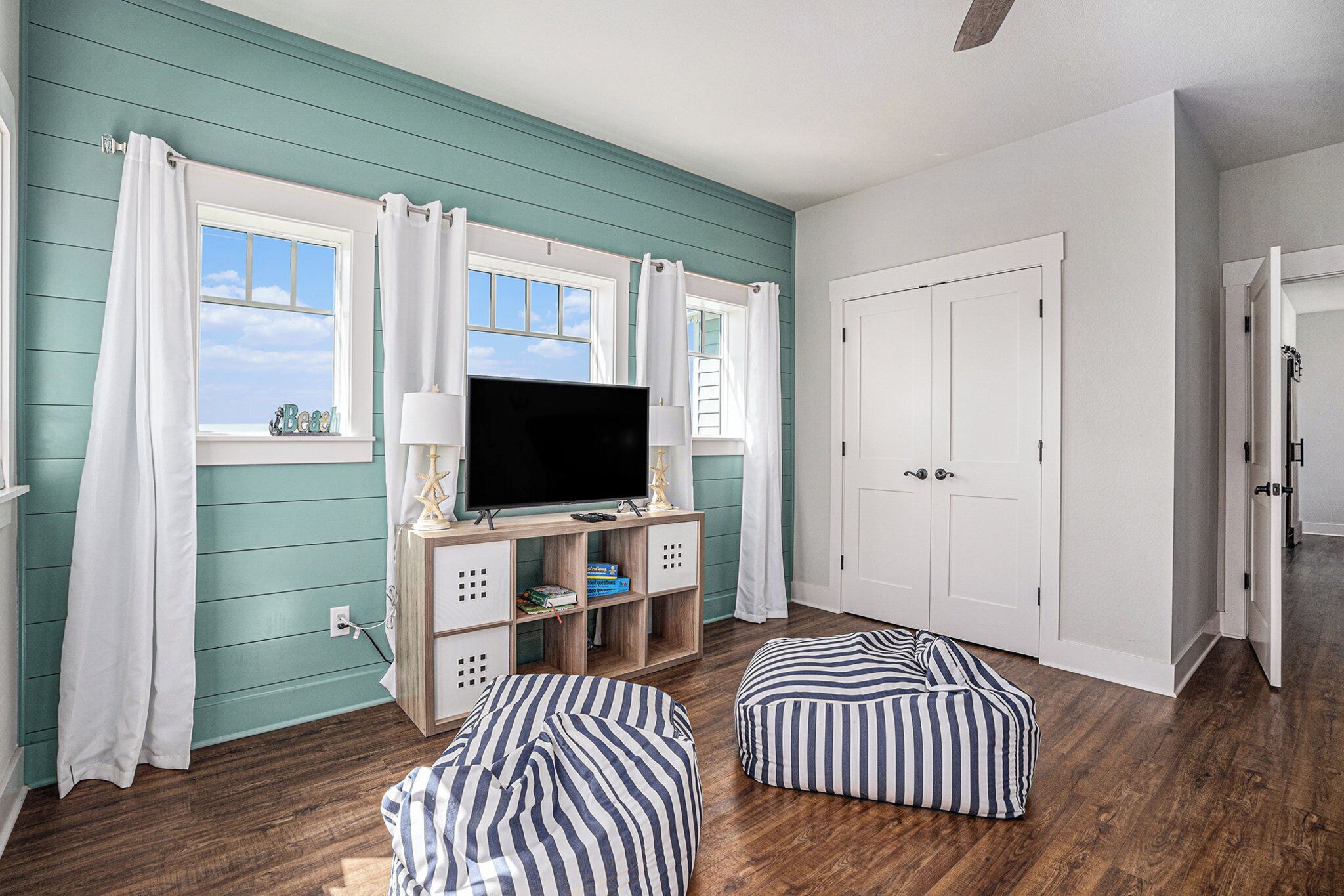 A room with teal walls, a television on a wooden stand, two striped ottomans, and a white door.