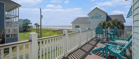 Une terrasse en bois avec une balustrade blanche, une maison dotée d’une grande fenêtre et une vue sur la plage.