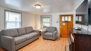 A room with a gray sofa, a smaller chair, a wooden desk, and a TV on a stand.