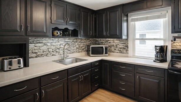 A modern kitchen with dark cabinets, a stone backsplash, a stainless steel sink, a microwave, a toaster, and a coffee maker.