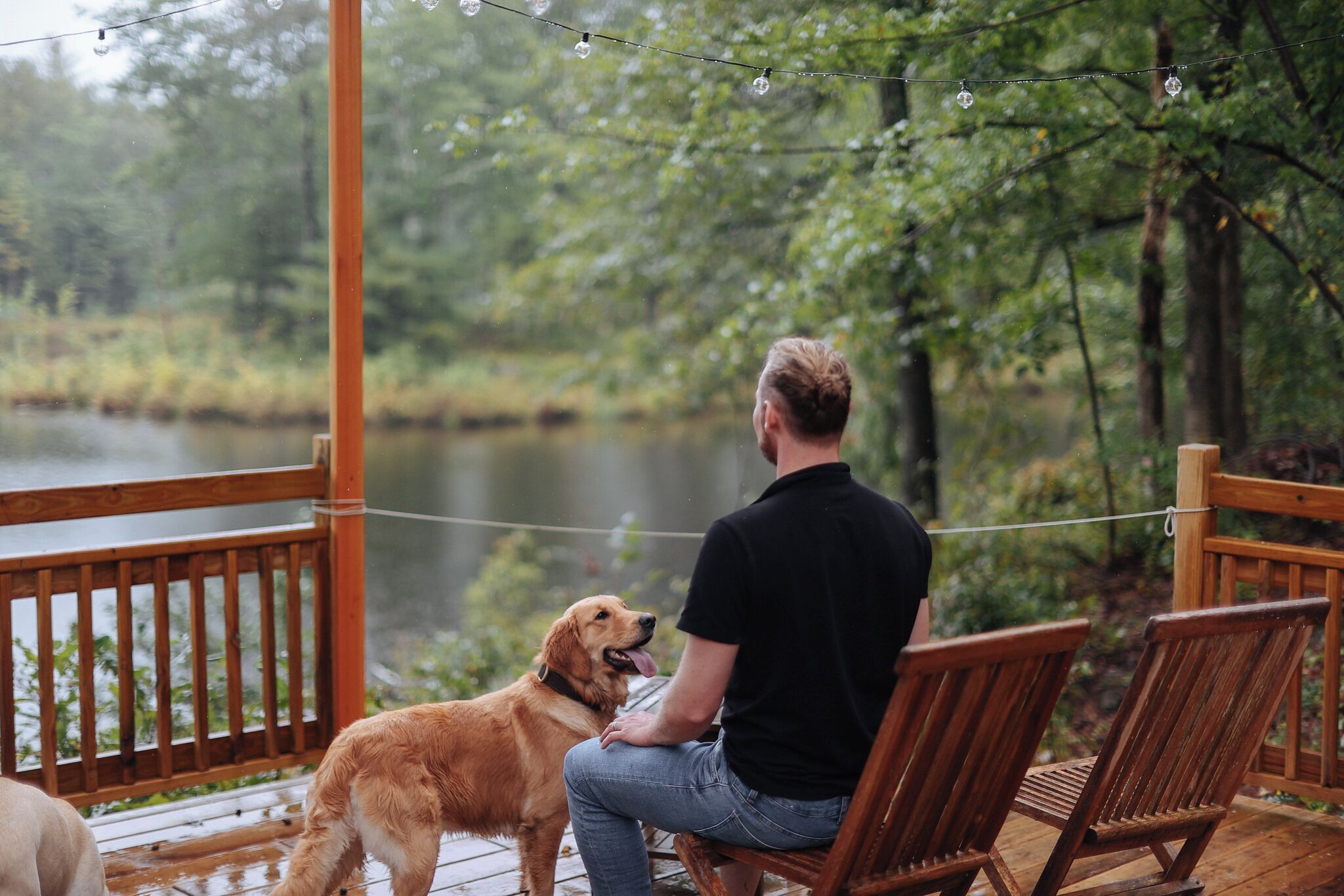 A man and a dog on a wooden deck with a pond and trees in the background.