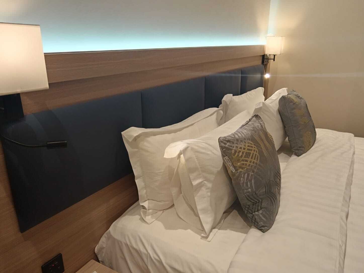 A hotel room with a bed, pillows, a bedside table, a lamp, and a wall-mounted light fixture.