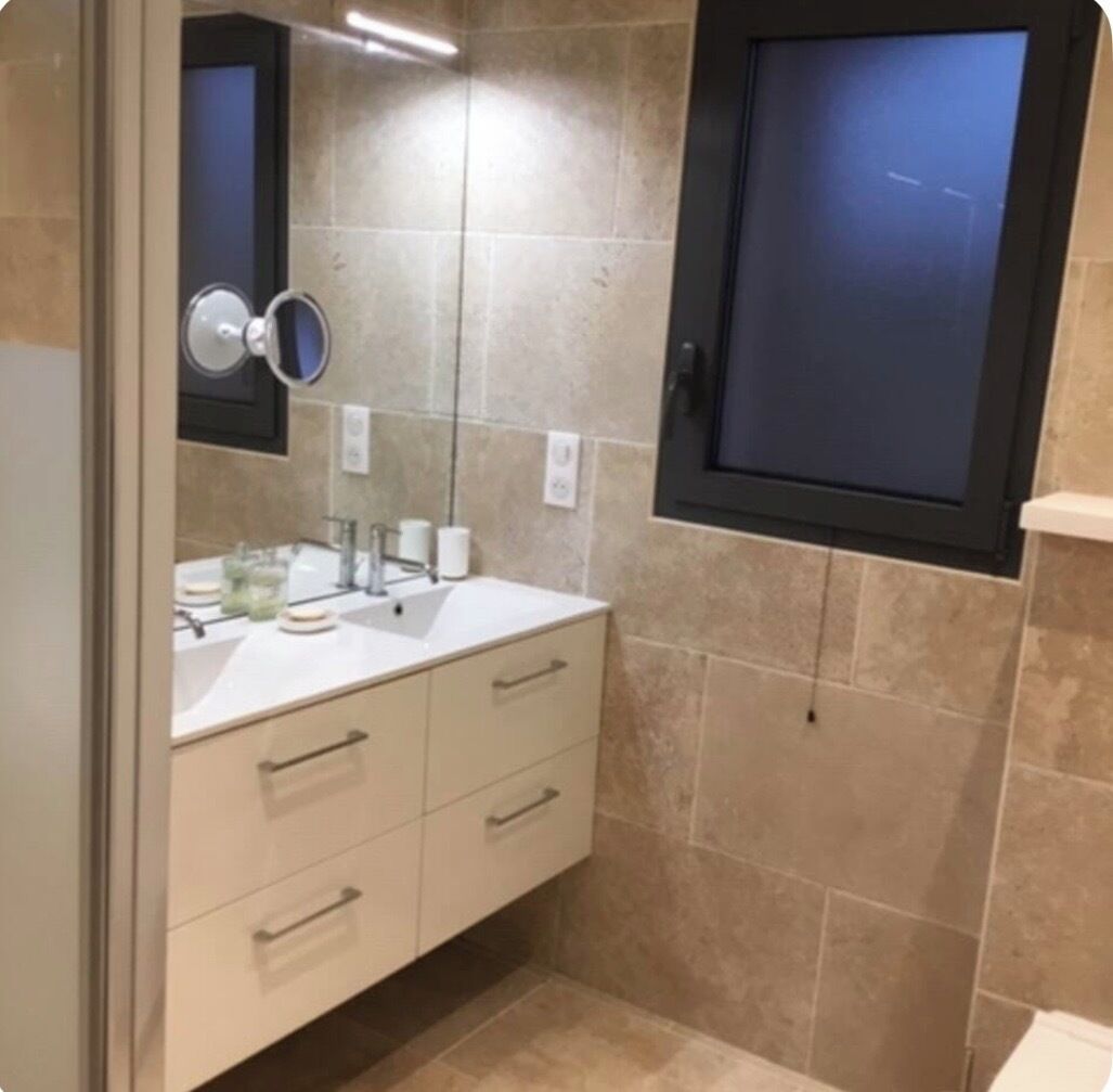 A bathroom with a white sink, mirror, and cabinet, tiled walls, and a window.