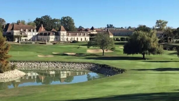 Ein Golfplatz mit Teich, Bäumen und Gebäuden im Hintergrund.