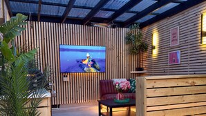 Un espace extérieur abrité, aménagé avec des places assises, un écran de télévision, des lattes en bois et un canapé rouge.