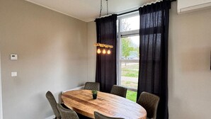 A dining room with a wooden table, six chairs, a hanging light fixture, and a view through large windows with black curtains. - Lodge 65 | 6 persoons chalet in Zennewijnen (Zennewijnen)