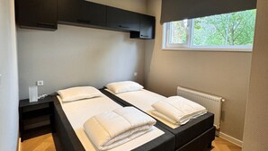 A bedroom with two beds, a window, a radiator, and a wall-mounted cabinet. - Lodge 65 | 6 persoons chalet in Zennewijnen (Zennewijnen)