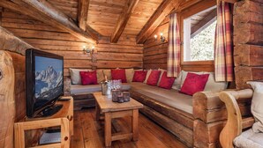 Interior de una cabaña de troncos con un sofá de madera, un televisor y una mesa con vasos y una vela.