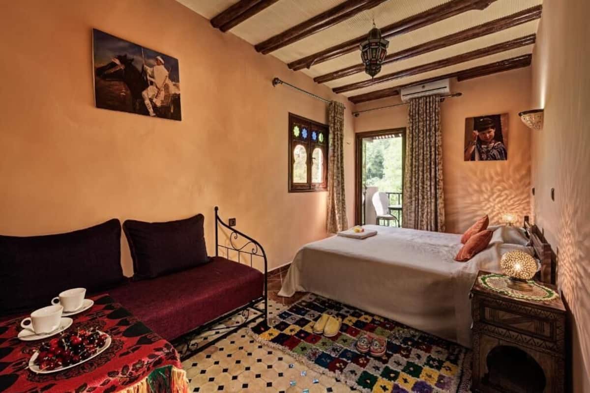 A cozy room with a bed, a sofa, a table with cups and a plate of fruit, and a patterned rug.