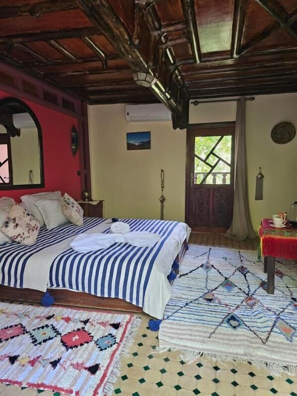 A room with a bed, a table with a red tablecloth, a patterned rug, and a door leading outside. - Riad Oussagou (Imlil)