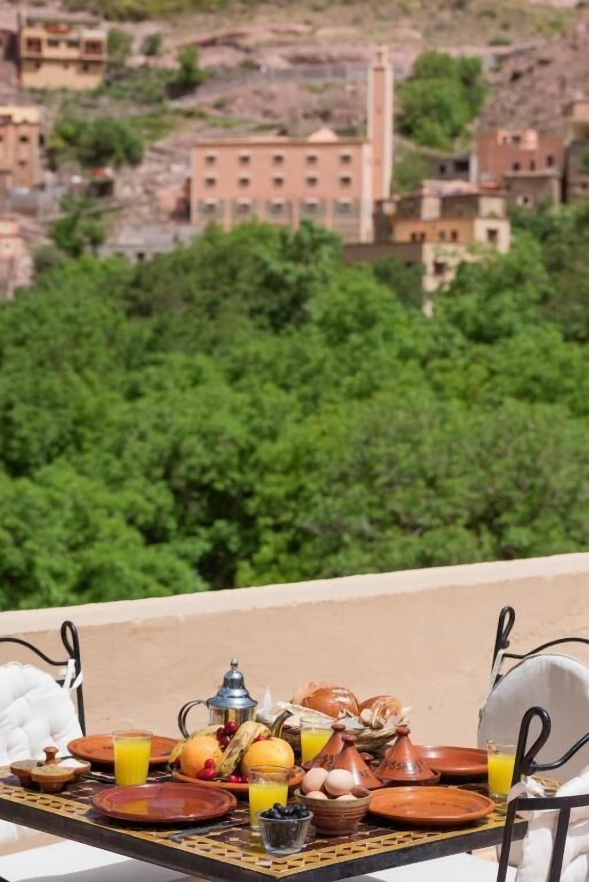 A table set with Moroccan breakfast items, including oranges, eggs, and a teapot, overlooking a mountainous landscape.