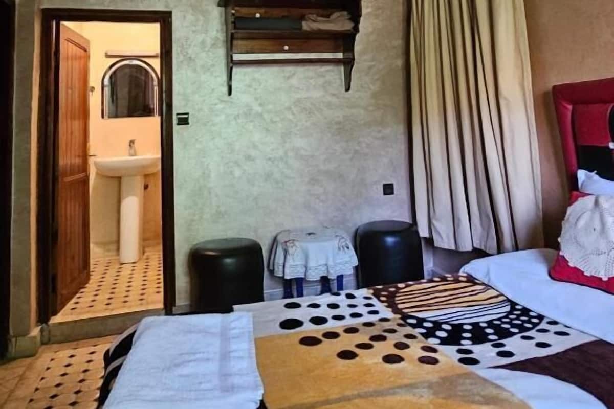 A hotel room with a bed, a small table, a mirror, a shelf, and a bathroom visible through an open door.