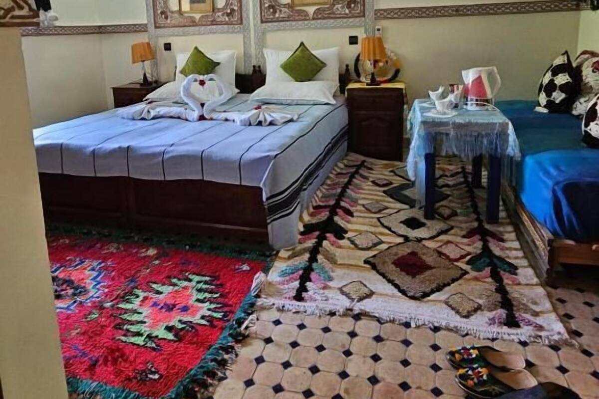 A hotel room with a bed, a small table with a lamp, a patterned rug, and a tiled floor.
