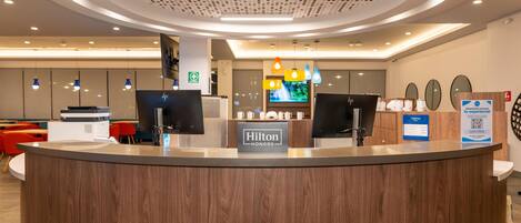 A hotel reception desk with a digital display, a sign for room rates, and a sign indicating a guest service area.