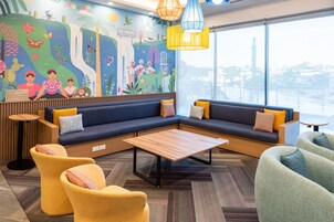 Un área de descanso moderna con un ventanal amplio, un mural que representa escenas culturales y una disposición de asientos que incluye sofás y sillones.