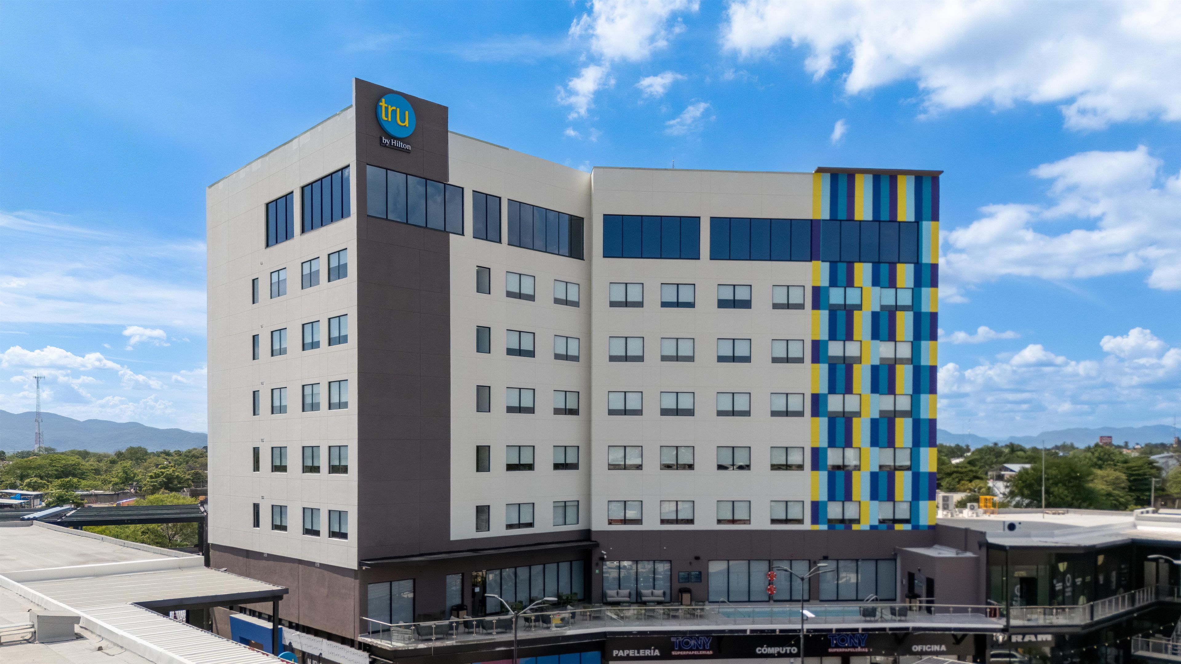 A modern hotel with a prominent 'tru by Hilton' sign, multiple floors, and a flat roof with a covered outdoor area.