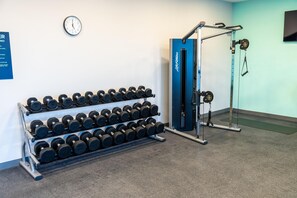 Een sportschool met een rek dumbbells, een krachttrainingapparaat en een klok aan de muur.