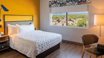A hotel room with a bed, a chair, a desk with a lamp, and a window with a view of a cityscape.