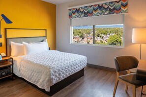 Une chambre d’hôtel avec un lit, une chaise, un bureau avec une lampe et une fenêtre donnant sur un paysage urbain.