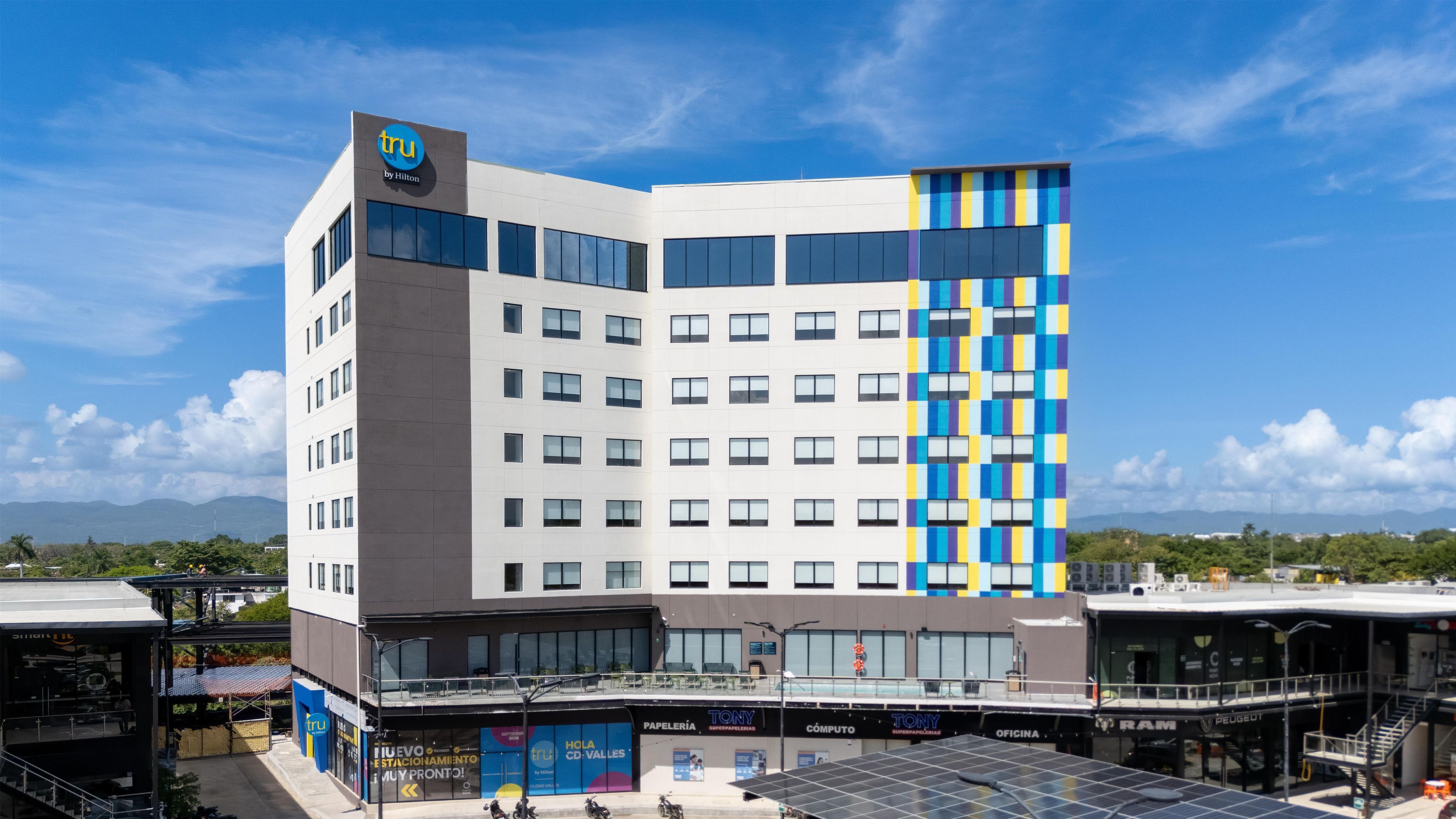 A hotel with a modern design, featuring a prominent logo at the top, multiple floors with large windows, and a ground-level commercial area with various shops.