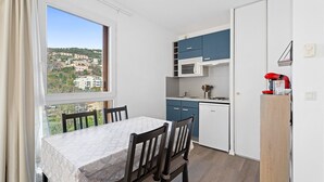 A compact kitchen with a dining table and chairs, a view of buildings through the window, and a microwave on the counter. - Air-conditioned flat with a balcony & swimming pool (Grasse)