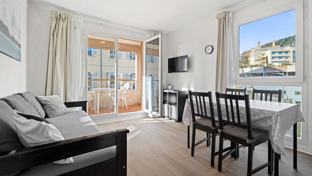 A modern living room with a sofa, dining table, chairs, a TV, and a balcony with a view of buildings. - Air-conditioned flat with a balcony & swimming pool (Grasse)