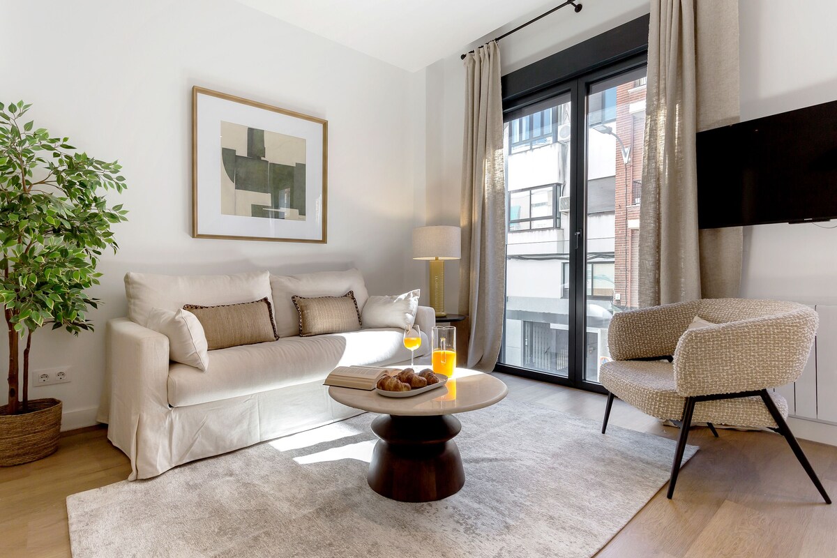 A modern living room with a sofa, a coffee table with croissants and juice, a floor lamp, a plant, and a TV on the wall.