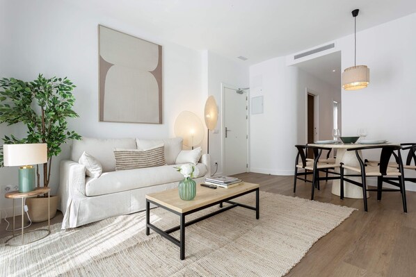 A modern living room with a white sofa, a wooden coffee table, a green vase, a lamp, a plant, and a dining area with a table and chairs. - Genteel Home Zabaleta (Madrid)