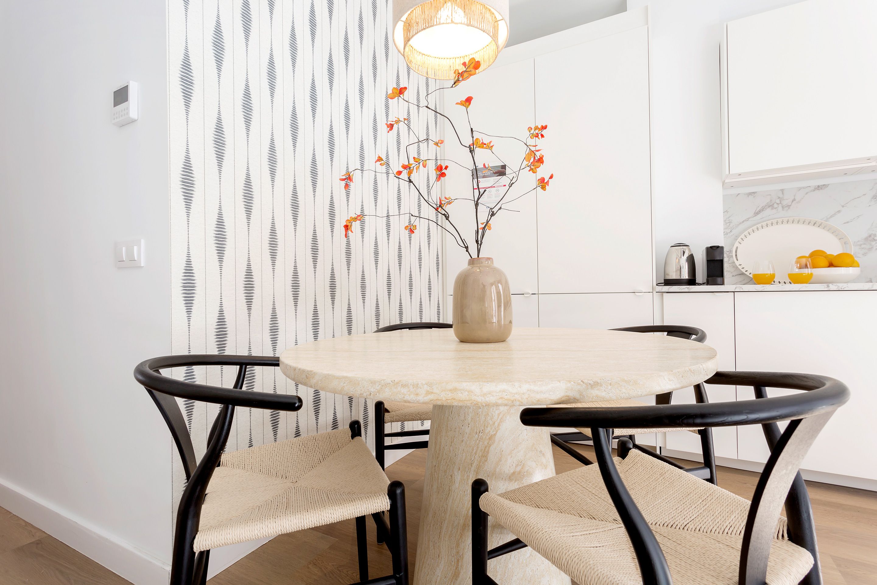 A modern kitchen with white cabinetry, a round dining table with woven chairs, and a vase with branches and leaves on the table.