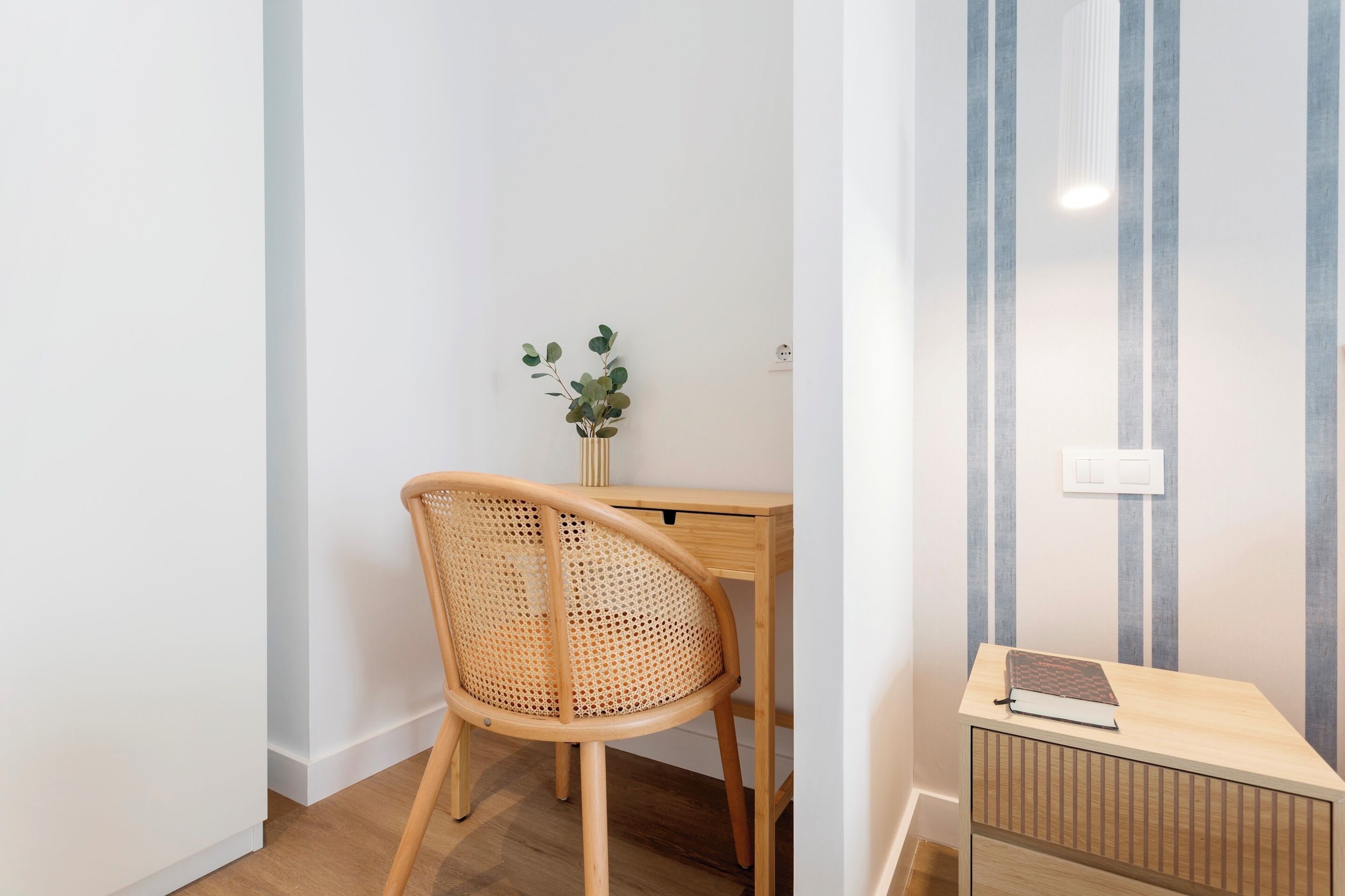 A small, neatly arranged workspace with a wooden desk, a wicker chair, and a small plant.