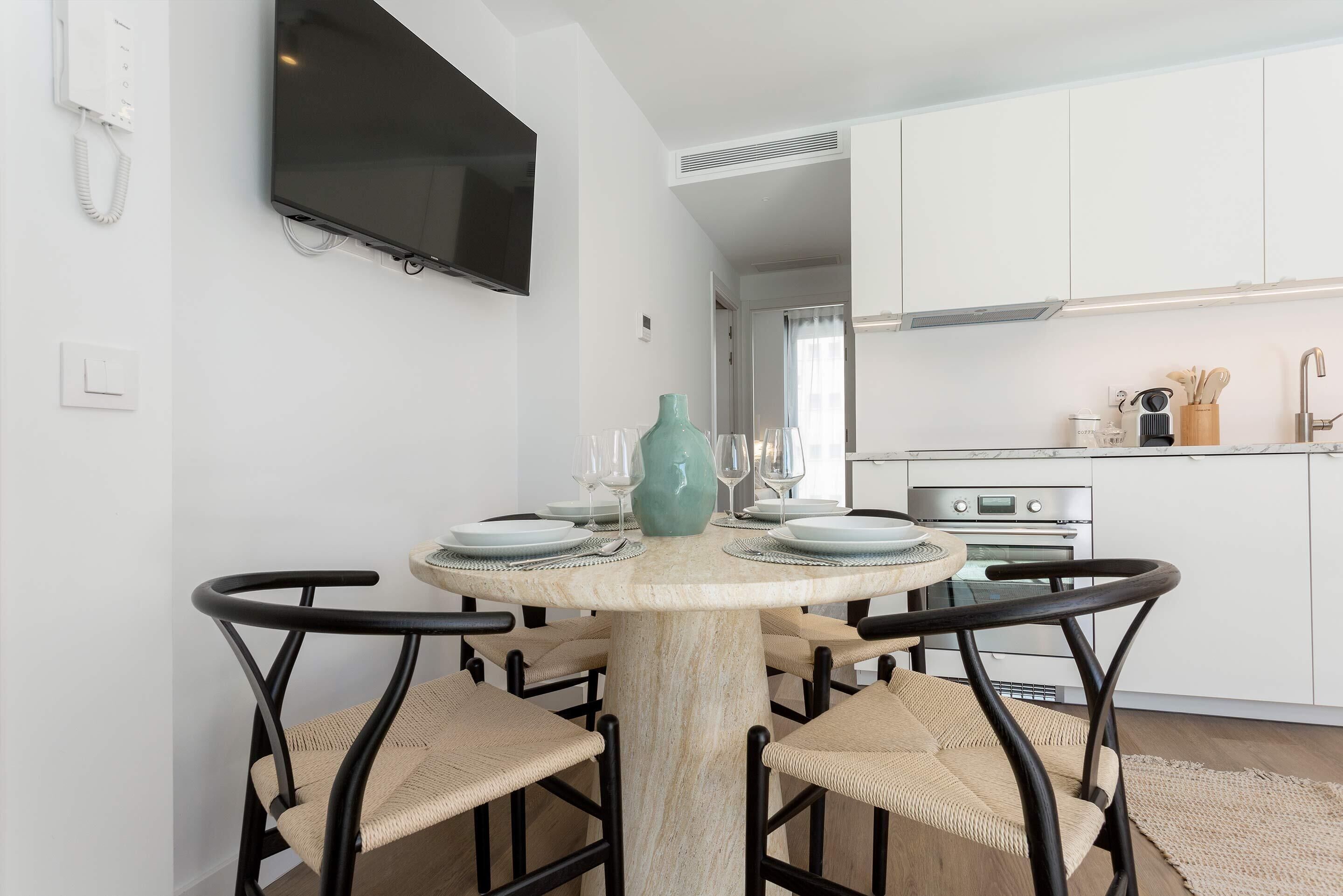 A dining area with a round table, chairs, a TV mounted on the wall, and a kitchen in the background.