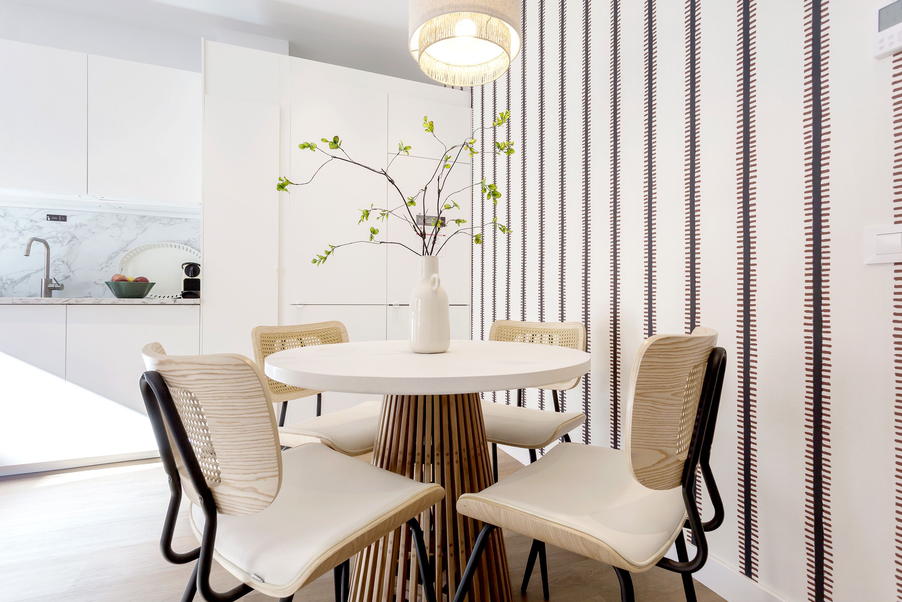 Un comedor moderno con una mesa redonda, cuatro sillas y un florero con ramas.