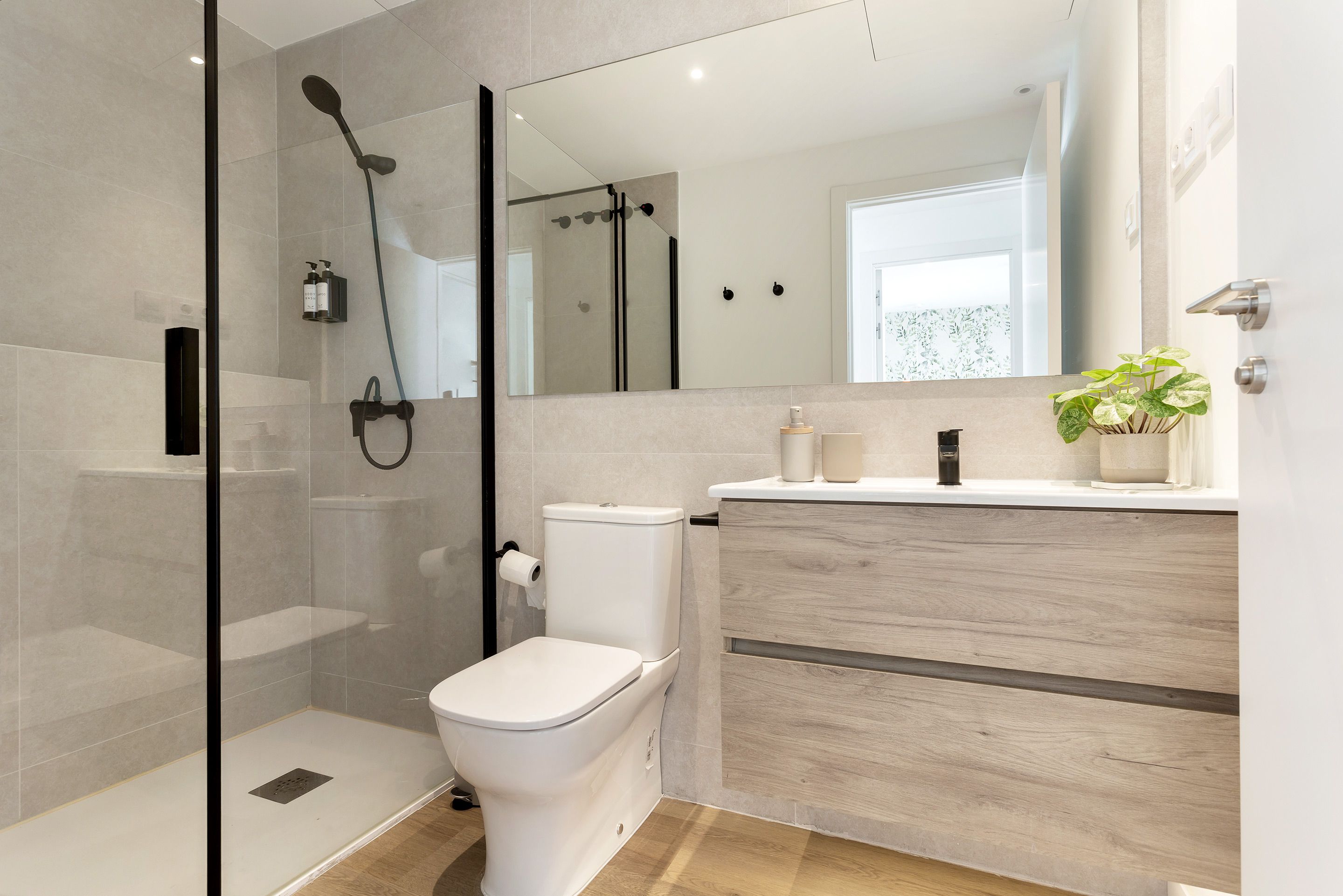Un baño moderno con ducha de vidrio, inodoro, tocador con espejo y una planta en maceta.