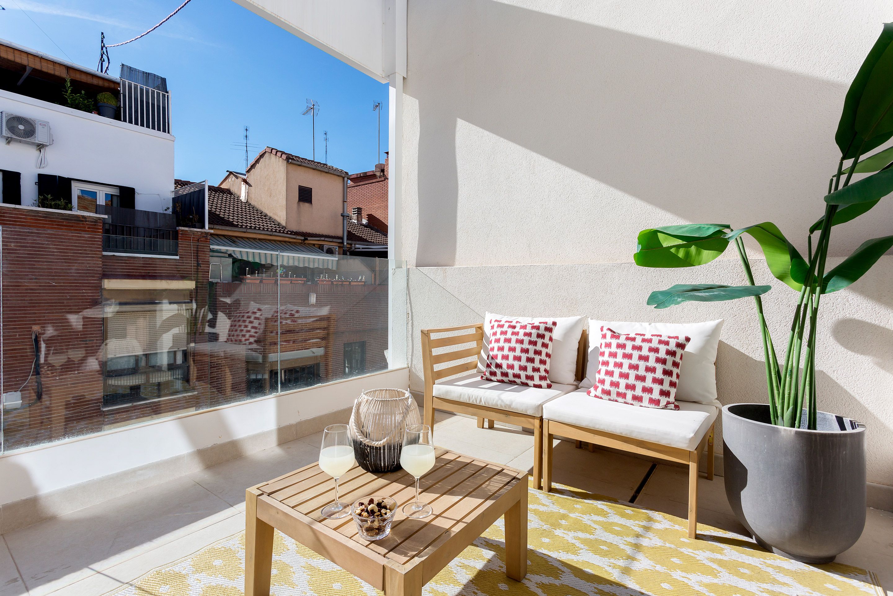 A balcony with a wooden table, two chairs, a potted plant, and a view of the neighborhood.