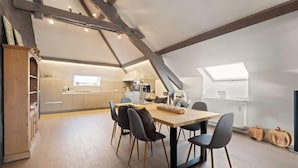 Een ruime loft met een hoge plafond, zichtbare balken, een eethoek met een tafel en stoelen, een keuken op de achtergrond en een dakraam.