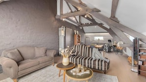 Een loft-achtige woonkamer met zichtbare houten balken, een bank, een gestreept fauteuil, een salontafel en een boekenkast.