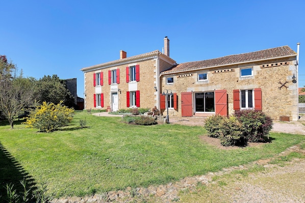A two-story stone building with red shutters, a grassy lawn, and a small garden area.