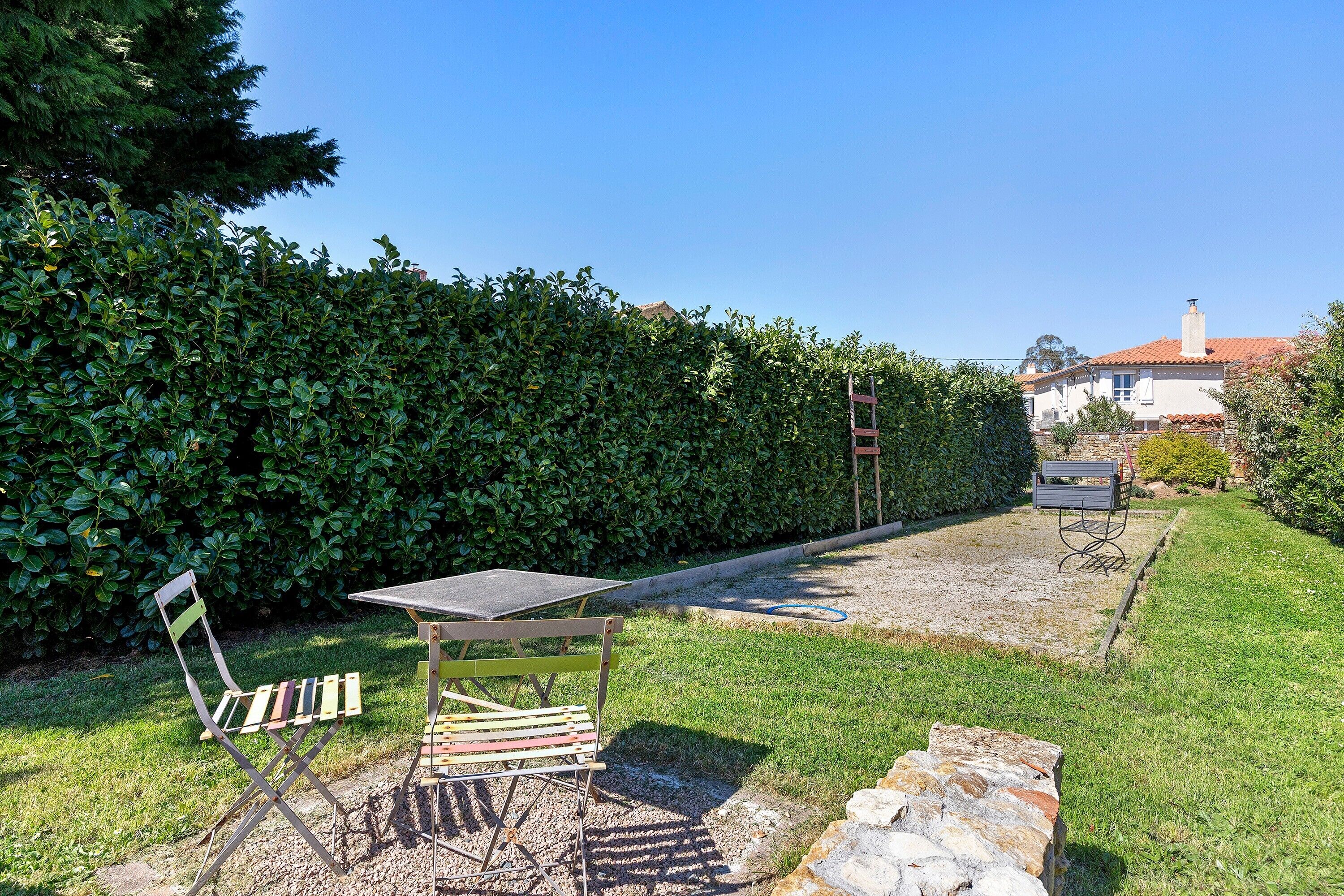 A garden with a table and chairs, a hedge, and a building in the background.