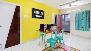 A hotel room with a yellow wall, a sign that reads 'HOTELO Company Serviced', a wooden desk with a printer, turquoise chairs, and a patterned rug.