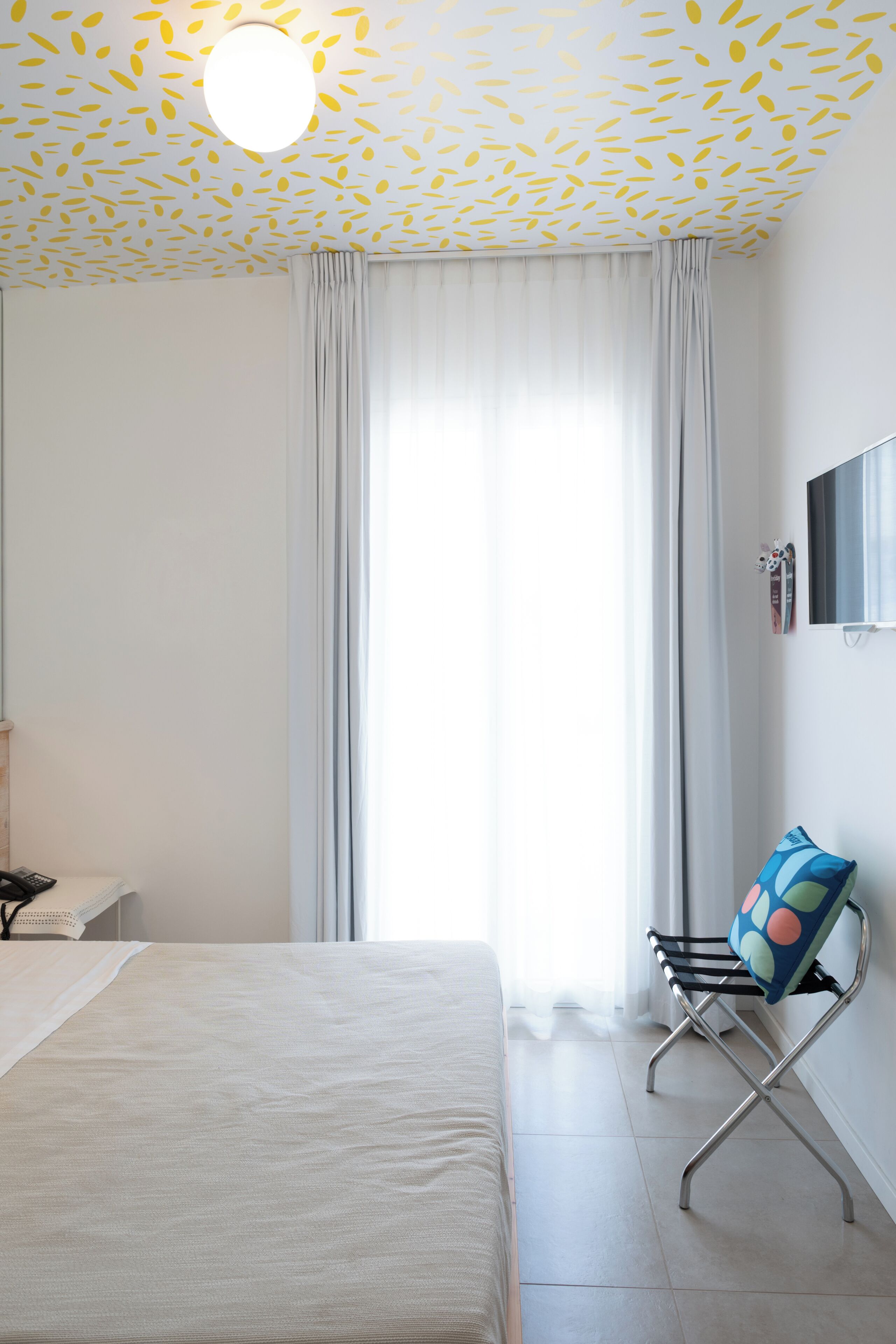 Een hotelkamer met een bed, een stoel met een kleurrijk kussen, een flatscreen televisie aan de muur en een plafond met een bladmotief.