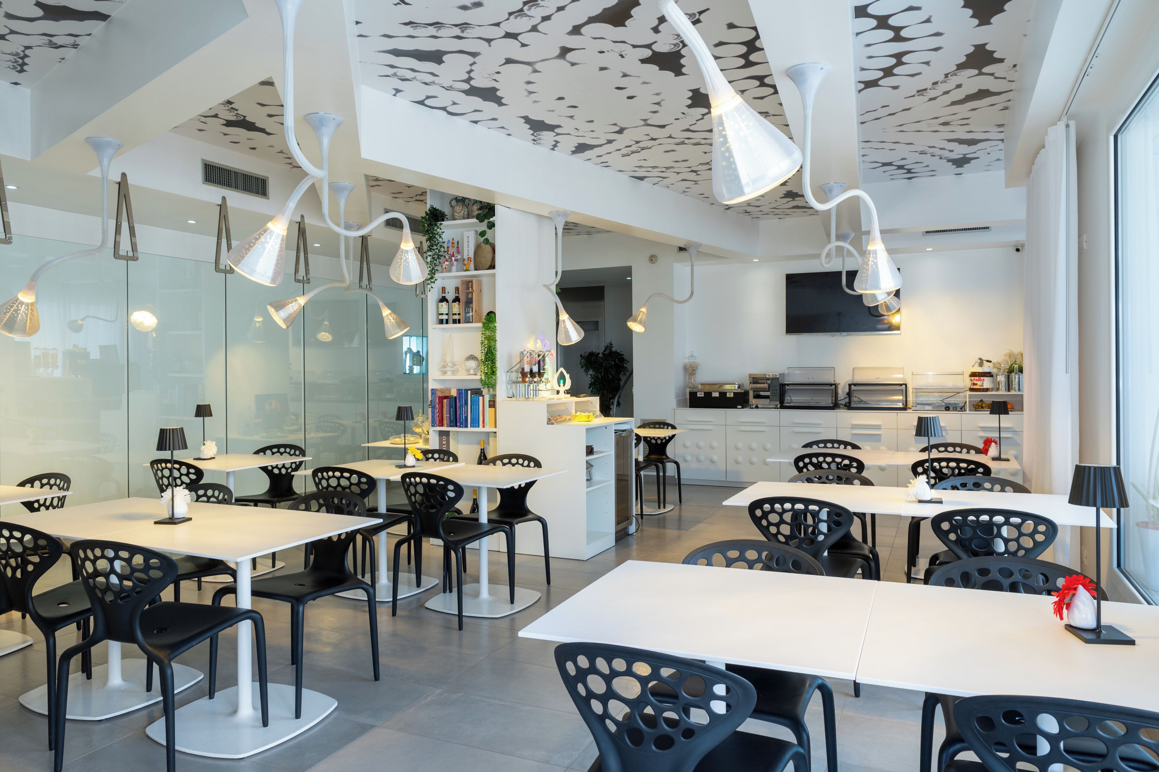 Un comedor moderno con mesas blancas y sillas negras, una cocina al fondo, e iluminación colgante.
