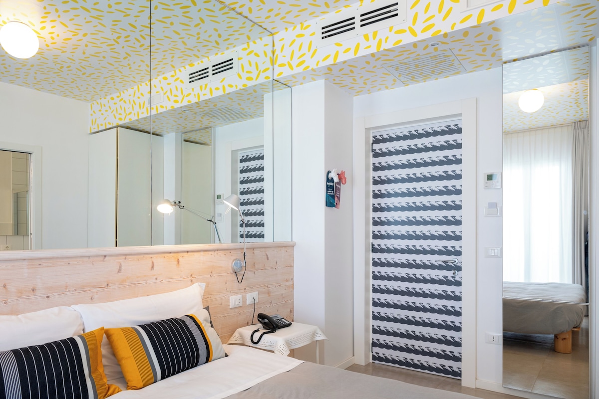 A hotel room with a wooden headboard, a bed with striped pillows, a bedside telephone, a mirror, and a patterned wall.