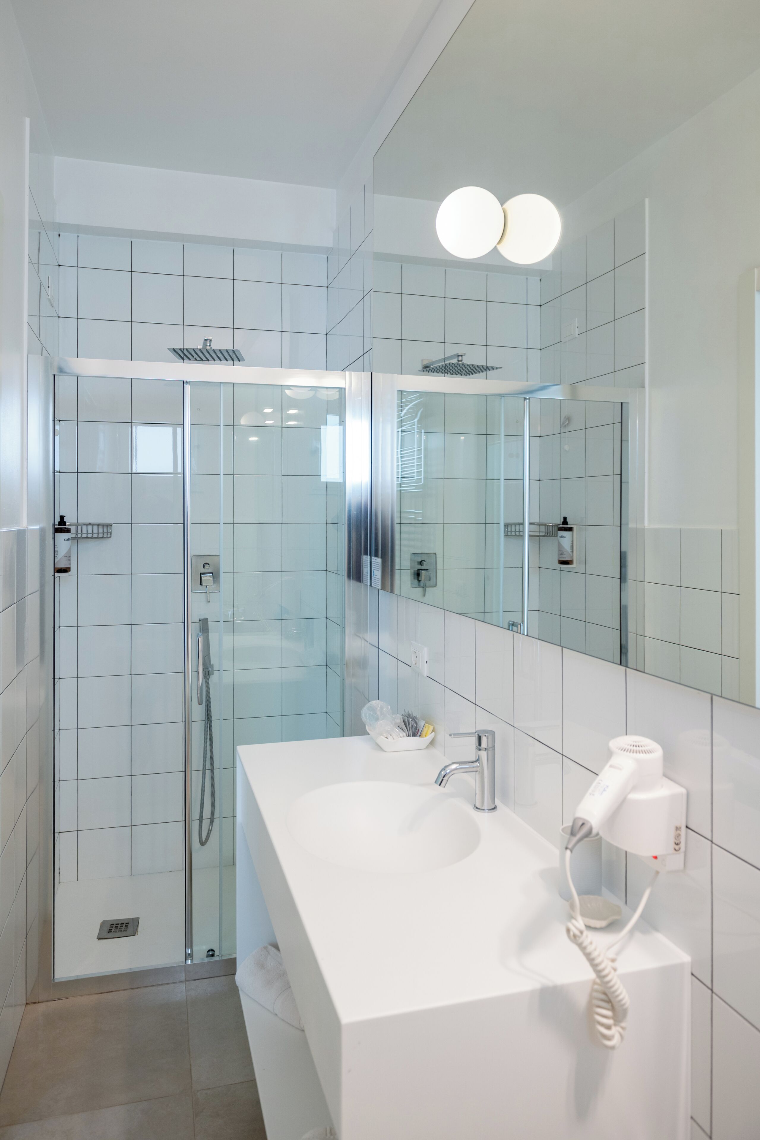 Un baño moderno con ducha envuelta en vidrio, tocador blanco con espejo y secador de pelo en la pared.