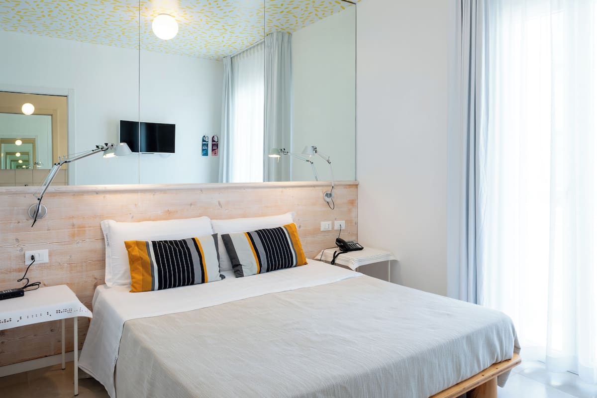 A modern hotel room with a wooden headboard, a large bed with white bedding, a bedside table with a lamp, a flat-screen TV, and a large mirror.