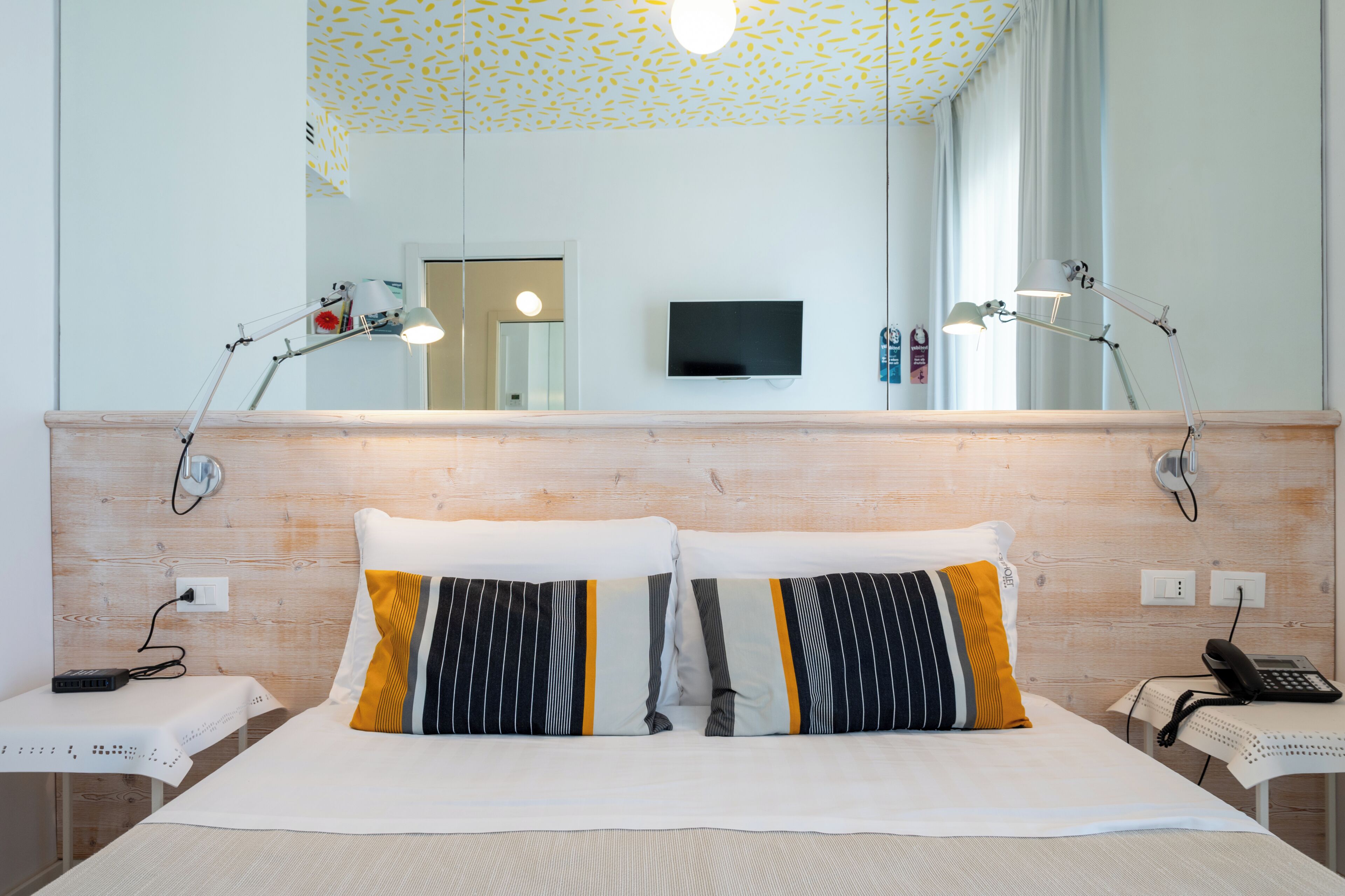 A hotel room with a wooden headboard, two bedside lamps, a flat-screen TV, and a telephone on a bedside table.