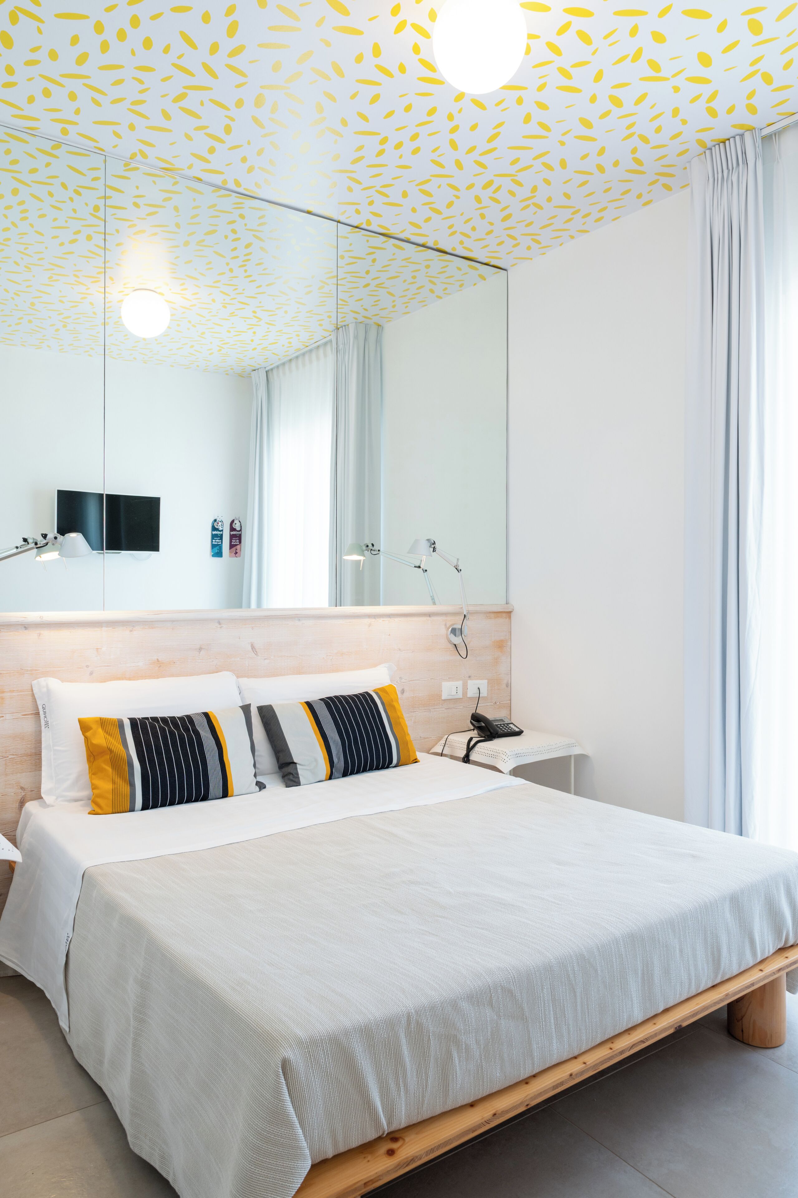 A modern hotel room with a large bed, a wooden headboard, a bedside table, a nightstand, a mirror, and a ceiling with a leaf pattern.