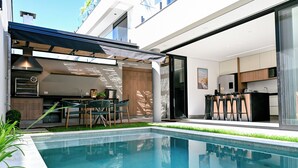Cocina exterior moderna con piscina, zona de comedor y puertas correderas de cristal que dan a un espacio de estar.